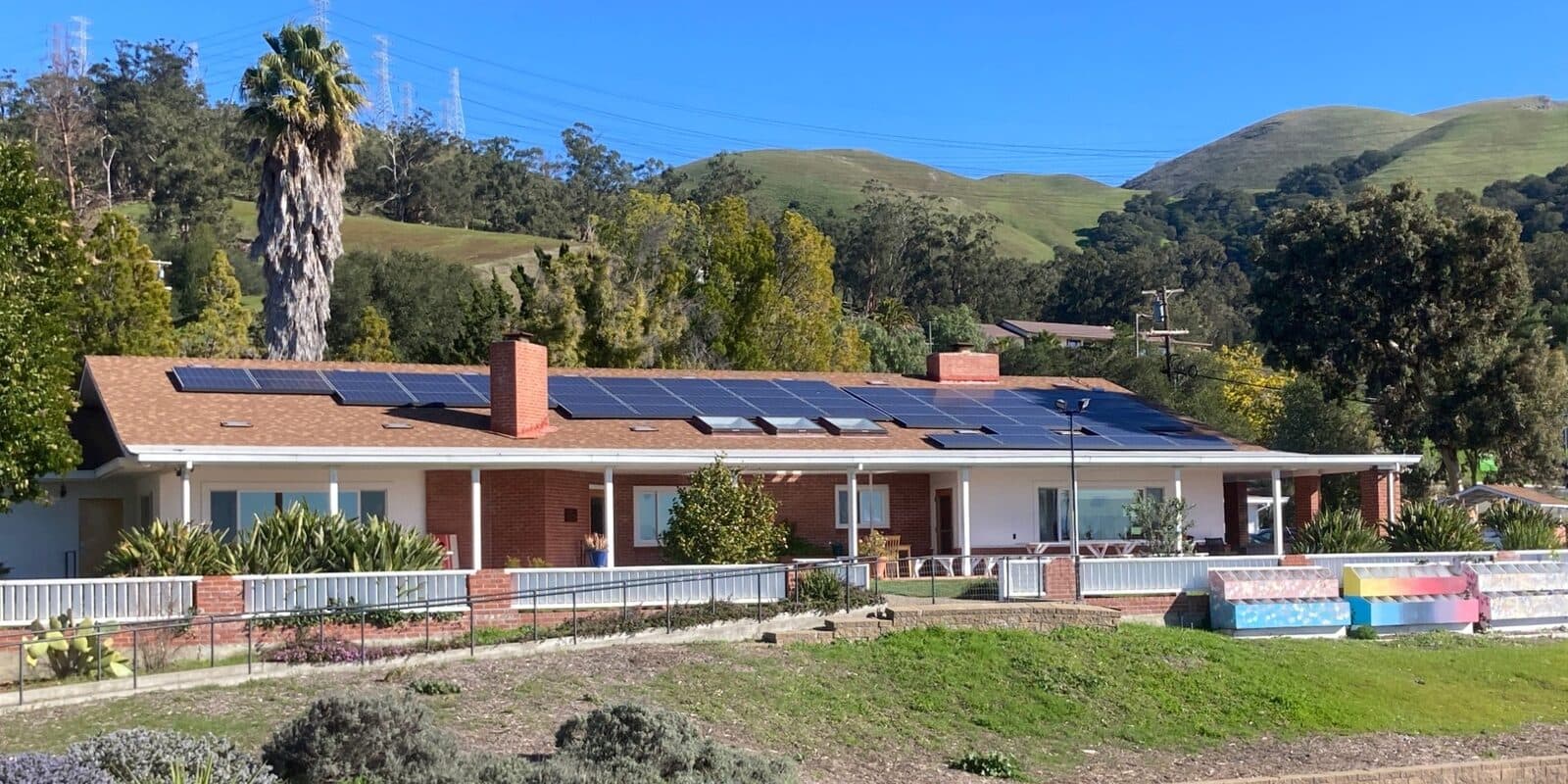 The upper campus building, a long brick and white ranch house with solar panels on its roof, seen across the playground with green hills behind