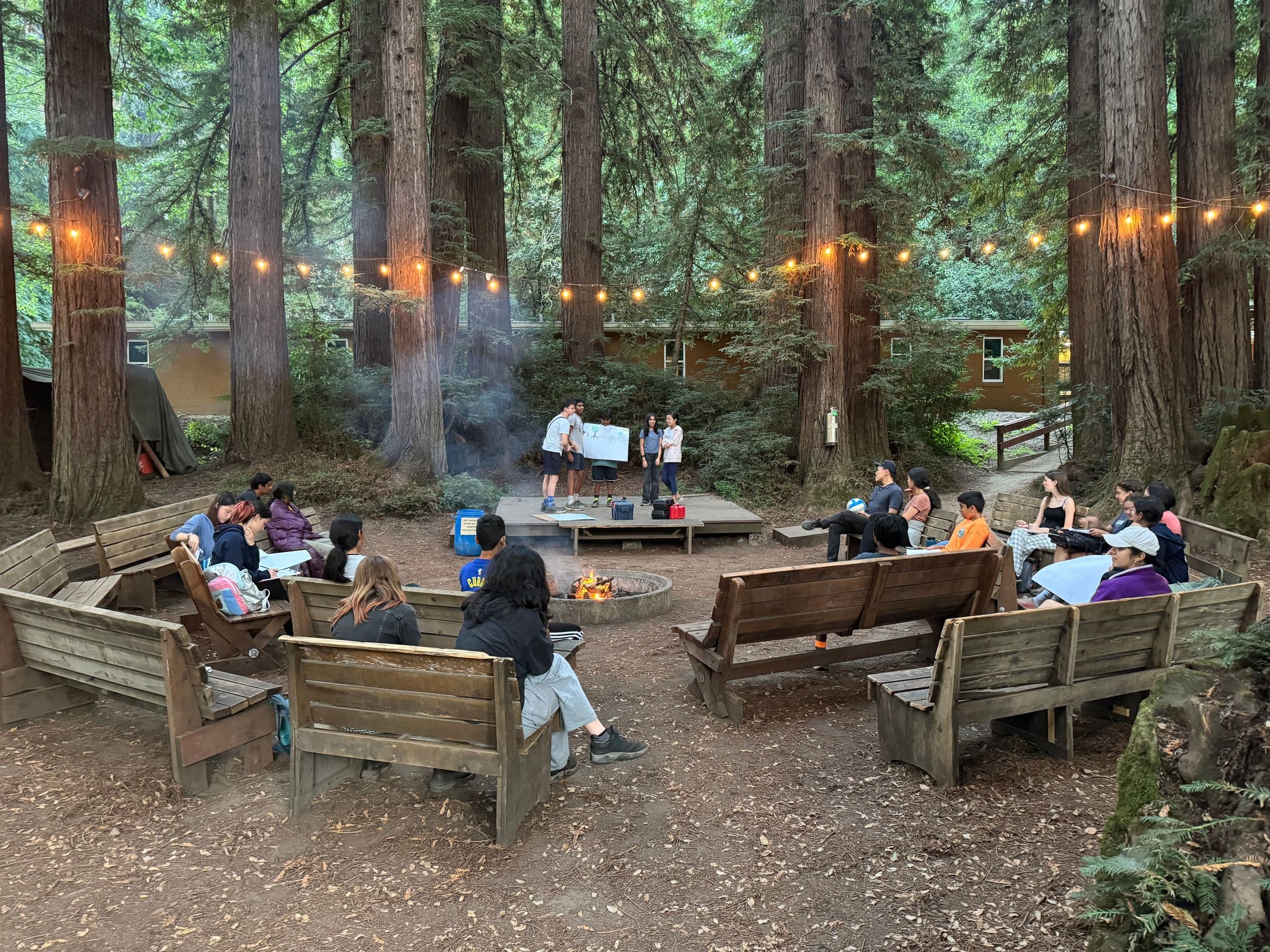 Students gathered on benches around a campfire among redwoods at School in the Woods